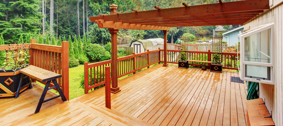 A spacious wooden deck with a pergola overlooks a lush garden. A bench and planters add charm, while an open window suggests fresh air and tranquility.